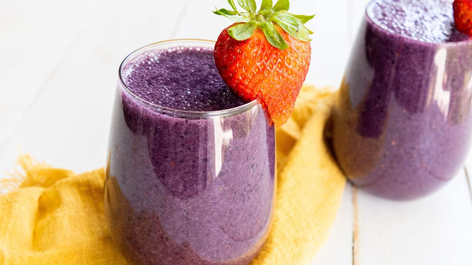 A glass of purple smoothie topped with a whole strawberry, placed on a white surface next to a yellow cloth.