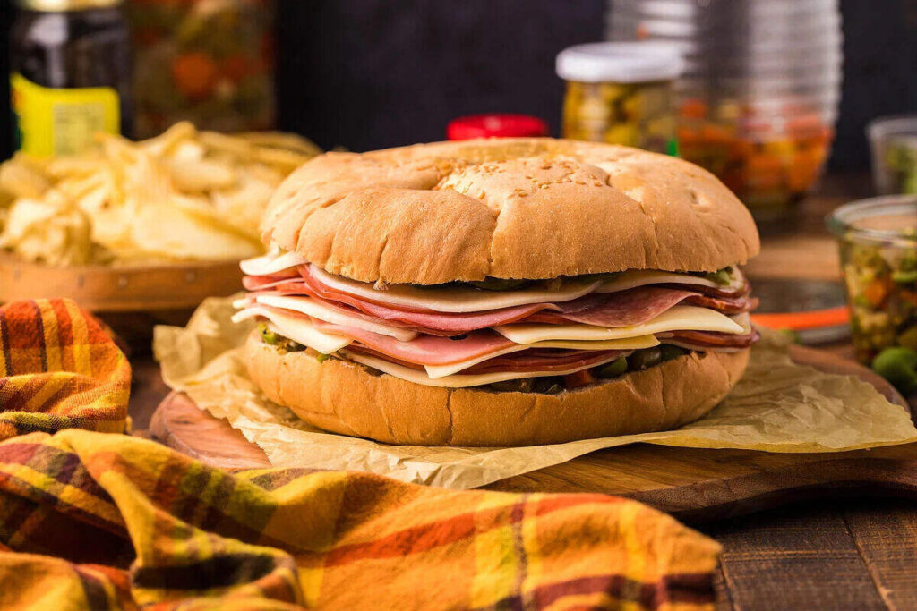 An easy sandwich favorite, this large stacked deli creation features layers of ham, salami, and cheese on a round bun, set on parchment paper with chips and jars of condiments in the background.