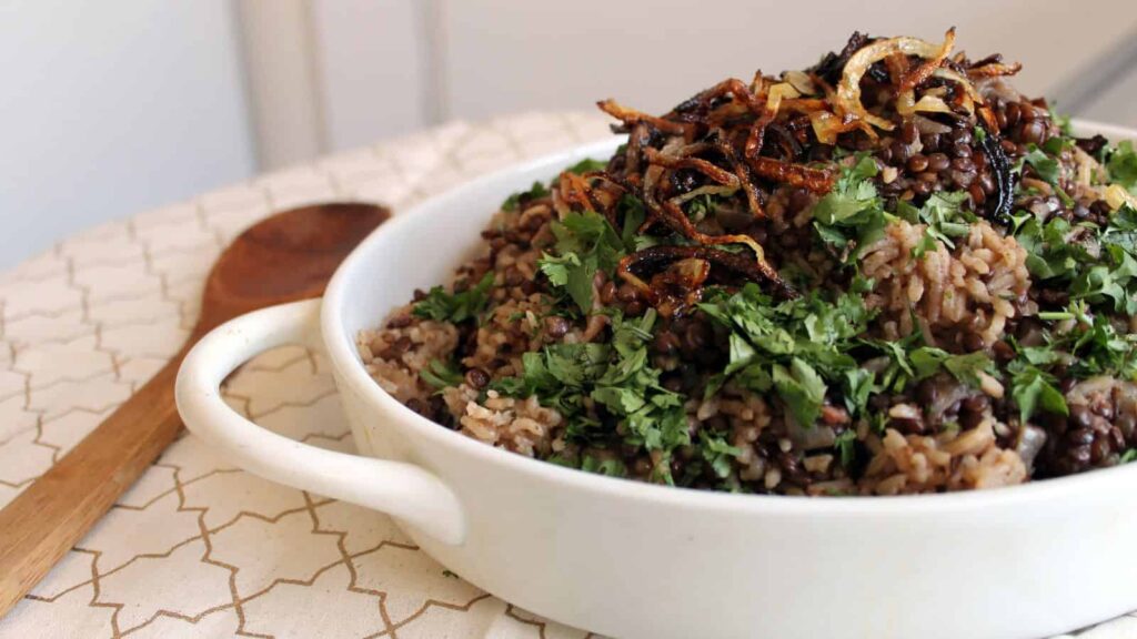 A white dish filled with Mujadara, a Middle Eastern rice and lentil dish, topped with fried onions and chopped parsley, sits on a patterned cloth beside a wooden spoon.