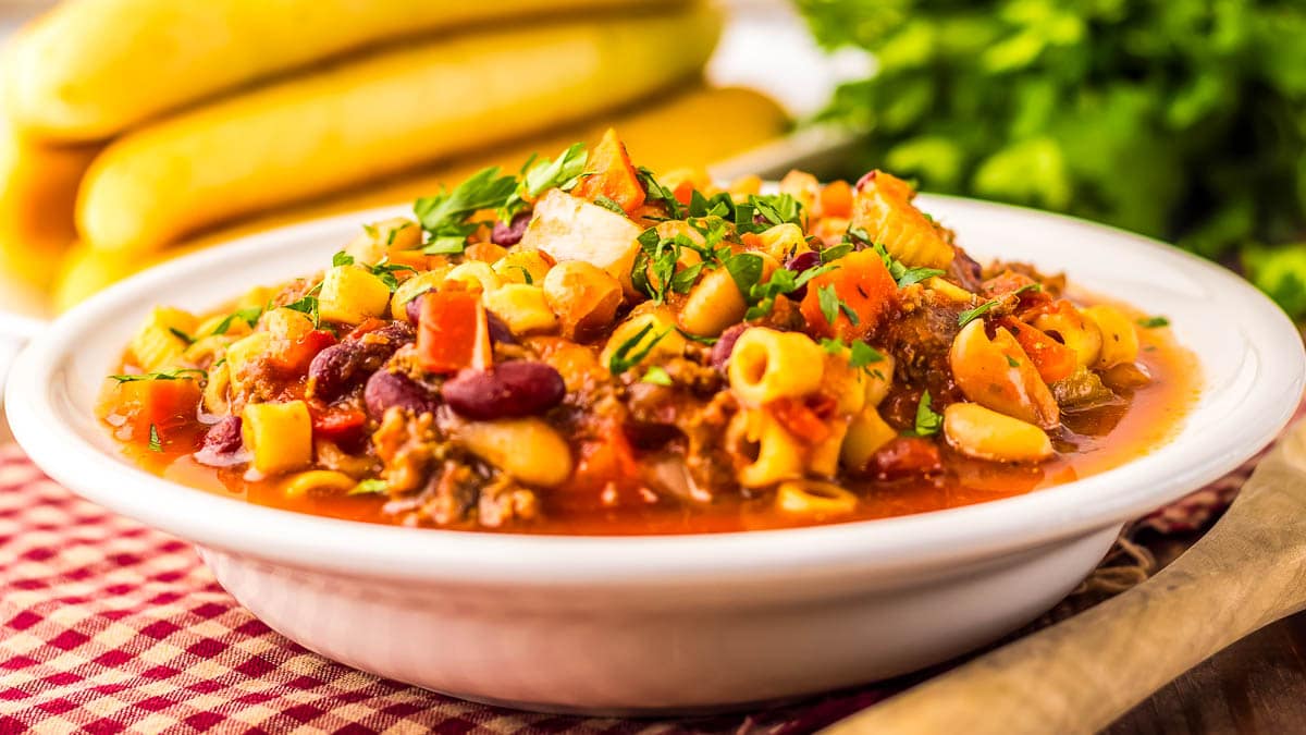A white bowl of soup with pasta, beans, vegetables, and herbs on a checkered cloth, with yellow squash and parsley in the background.