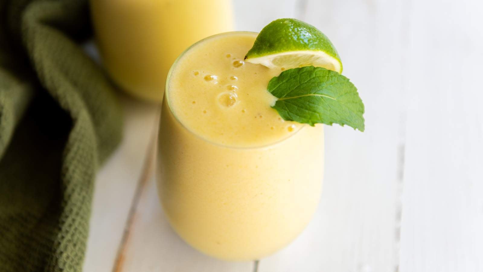 A glass of pineapple and banana smoothie garnished with a lime wedge and a mint leaf, placed on a white wooden surface.