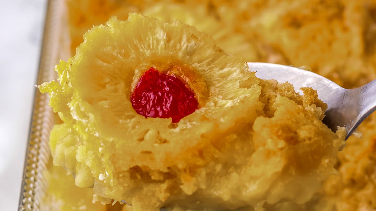 A close-up of a serving of pineapple upside-down cake on a silver spoon, showing a slice of pineapple with a red cherry in the center.