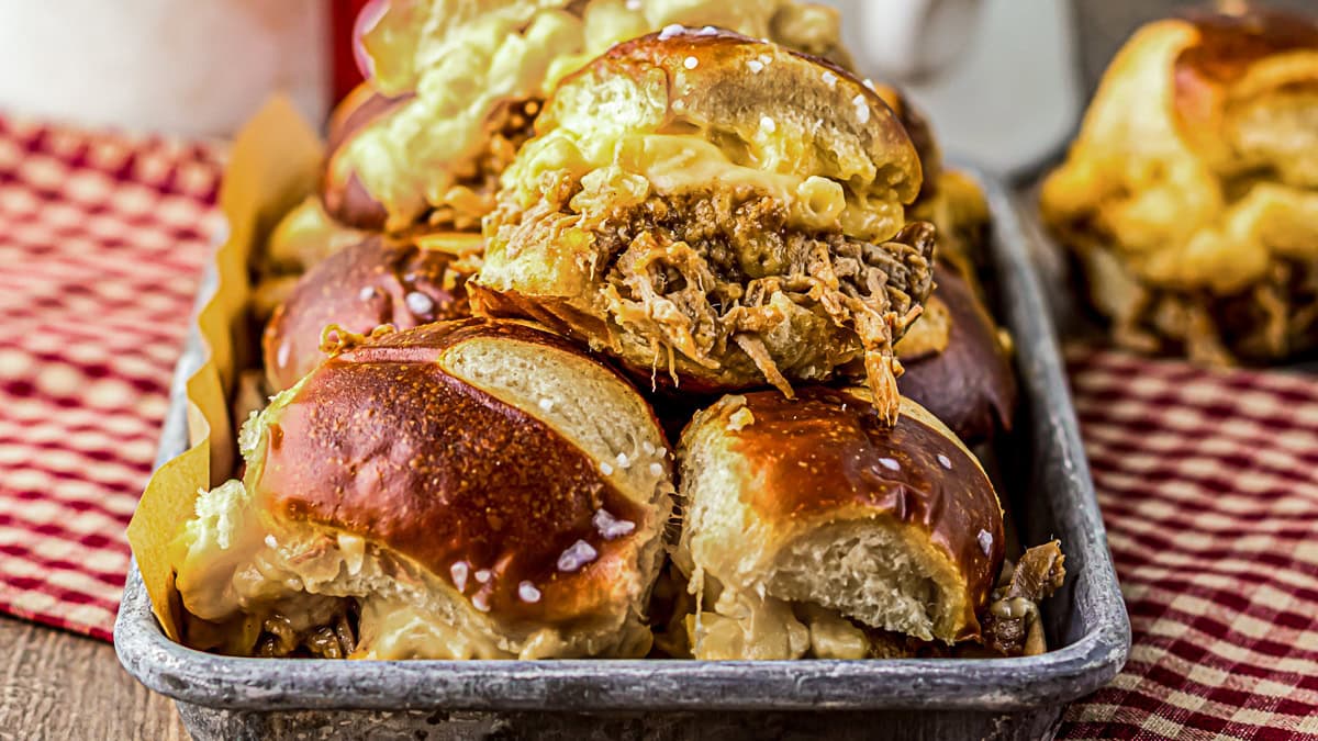 A tray of pretzel buns filled with shredded meat and melted cheese, arranged closely together on a red checkered cloth.