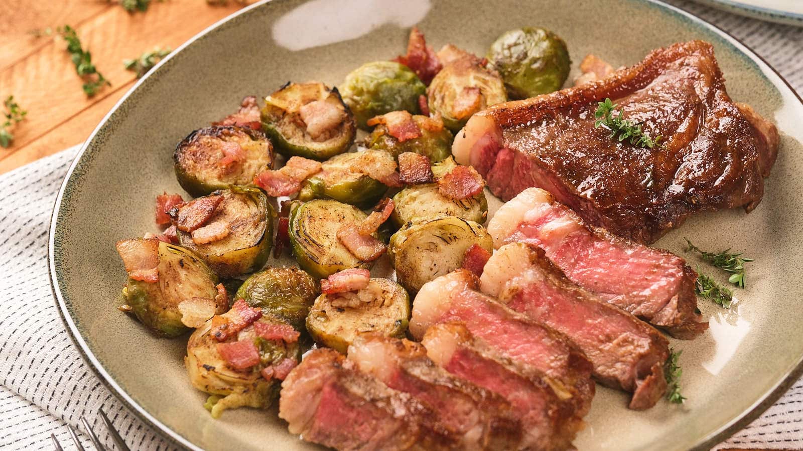 A plate with sliced steak and roasted Brussels sprouts mixed with bacon pieces, garnished with a small sprig of herbs.