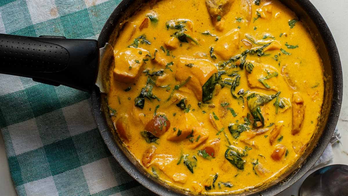 A pan of creamy curry with chunks of vegetables and greens, resting on a checkered cloth.