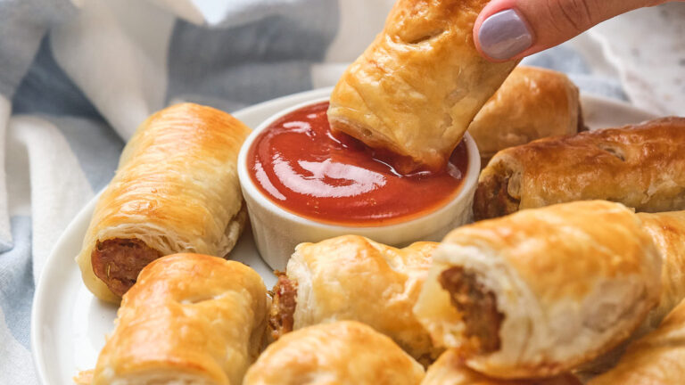 A hand dips a sausage roll into a bowl of tomato sauce.