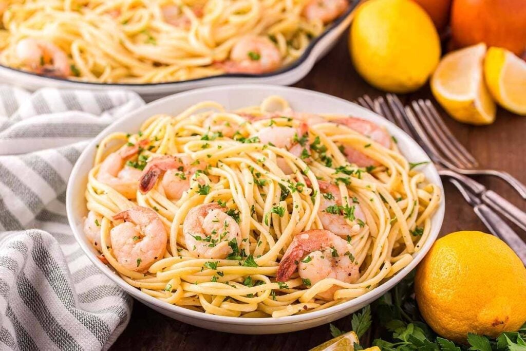 A bowl of shrimp pasta garnished with chopped herbs, surrounded by lemons, utensils, and a striped napkin on a wooden table.