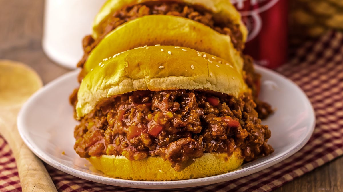 Three sloppy joe sandwiches filled with seasoned ground beef and diced red peppers on a white plate, placed on a red and white checkered cloth.