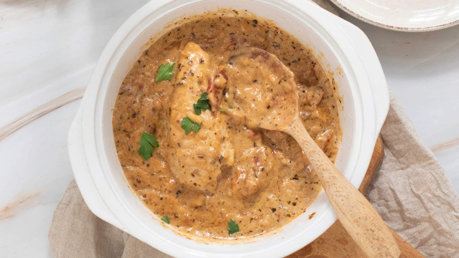 A bowl of cooked chicken garnished with fresh parsley, placed on wooden board and a wooden spoon inside.