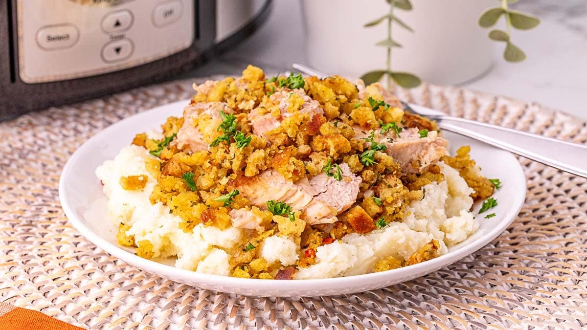 A plate of mashed potatoes topped with sliced turkey and stuffing, garnished with chopped parsley, on a woven placemat with a fork beside it.