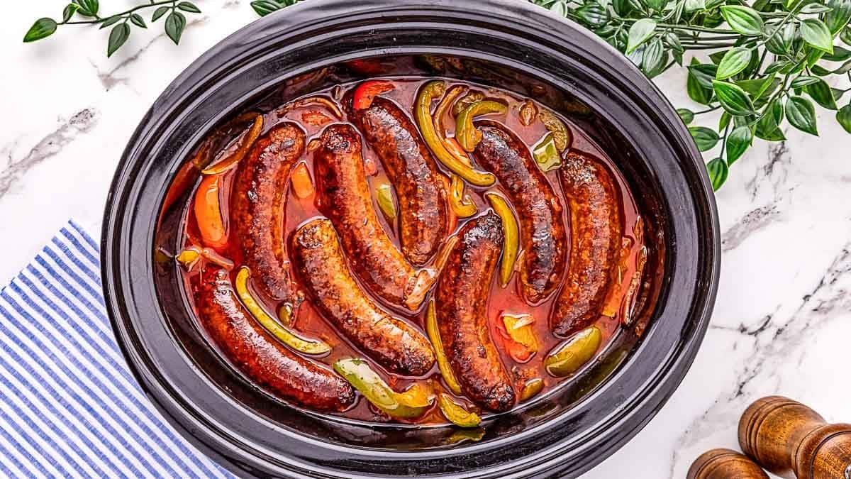 A slow cooker filled with cooked sausages, sliced bell peppers, and tomato sauce, with a blue-striped cloth and green leafy plant nearby.