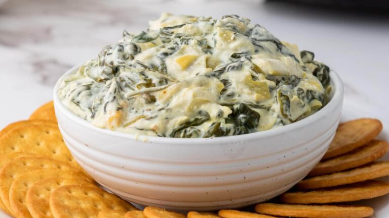 A white bowl filled with creamy spinach artichoke dip is surrounded by round crackers on a white surface.
