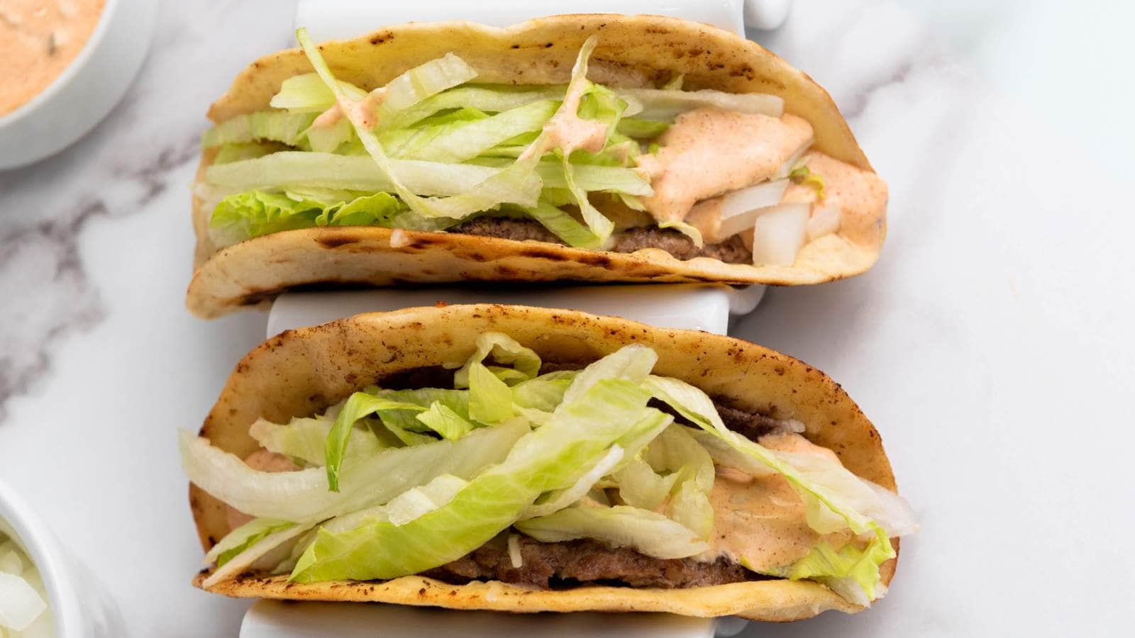 Two smash burger taco in taco holders topped with shredded lettuce, diced onions, and a creamy sauce.