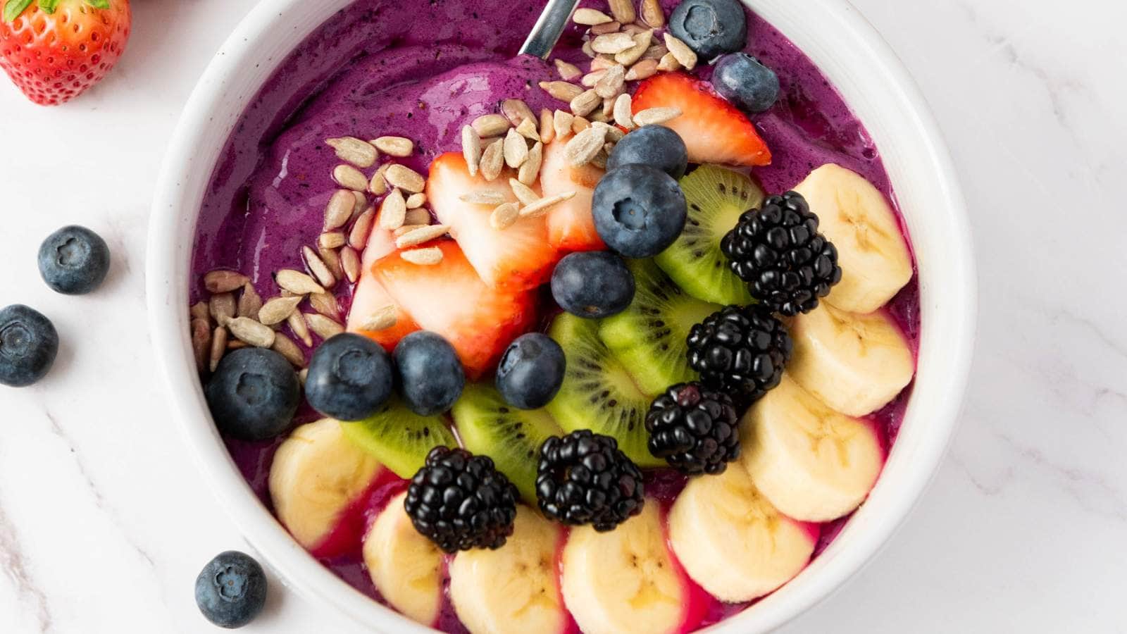 A smoothie bowl topped with sliced strawberries, bananas, kiwi, blueberries, blackberries, and sunflower seeds.