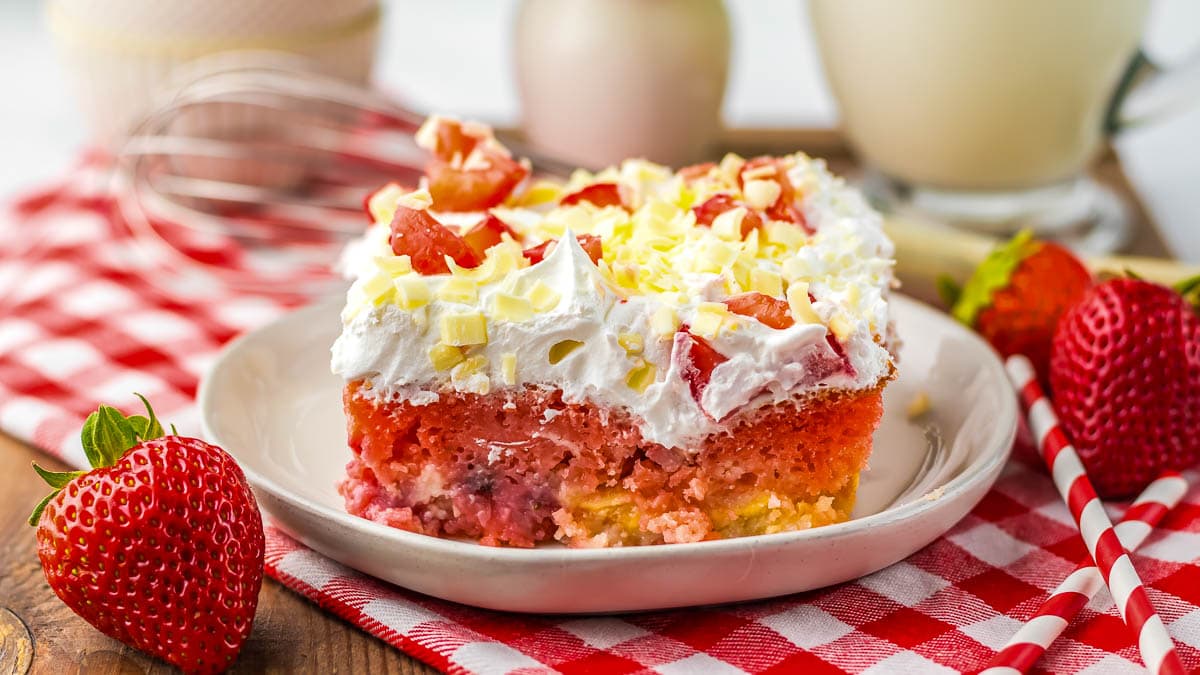 A slice of strawberry poke cake with white frosting and white chocolate shavings on a white plate. The plate is placed on a red and white checkered cloth.