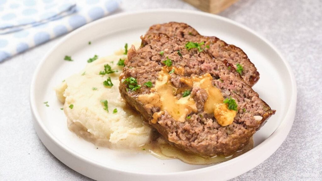 A plate with two slices of meatloaf topped with gravy, served alongside a portion of mashed potatoes.