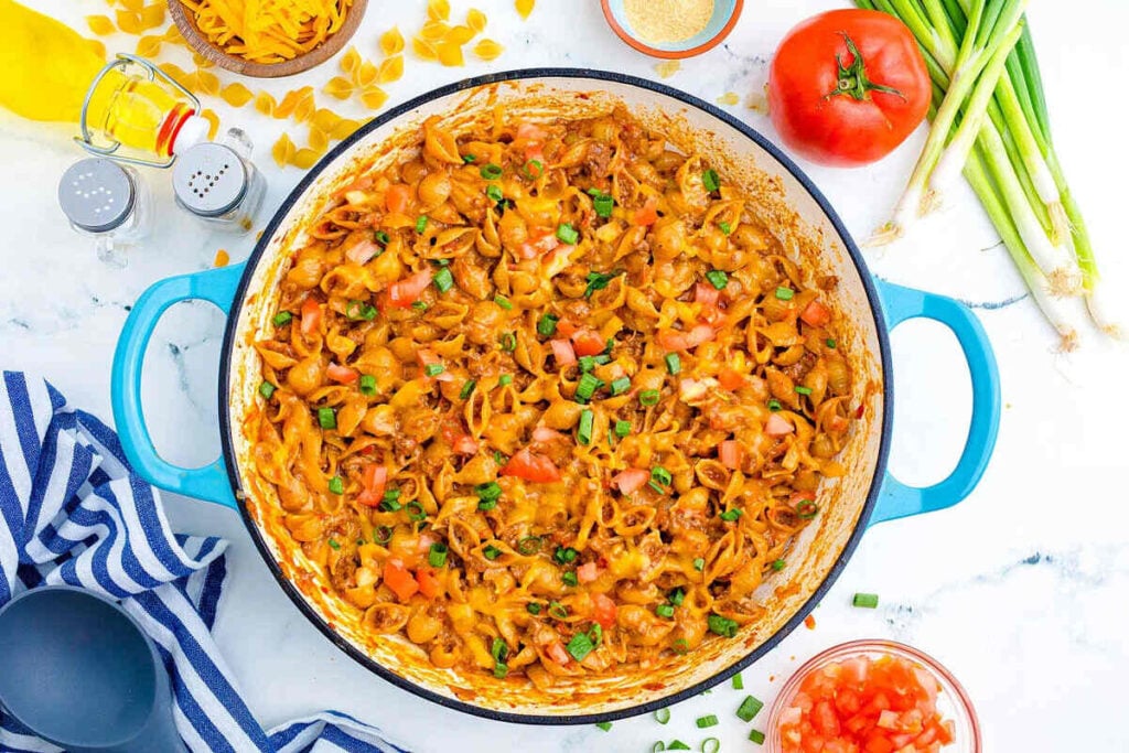 A large blue pot filled with cheesy pasta shells, diced tomatoes, and chopped green onions sits on a marble countertop with fresh vegetables and kitchen items nearby.