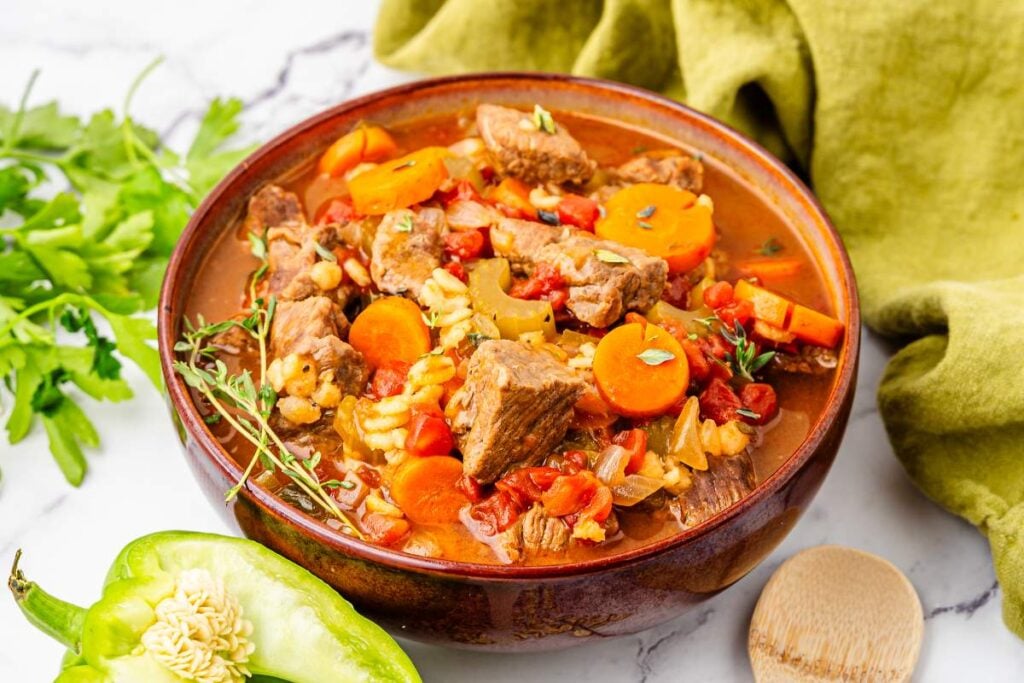 A bowl of beef stew with chunks of beef, sliced carrots, tomatoes, celery, and herbs, garnished with fresh parsley—an inviting choice for those seeking flavourful beef recipes—served beside a wooden spoon and green pepper.