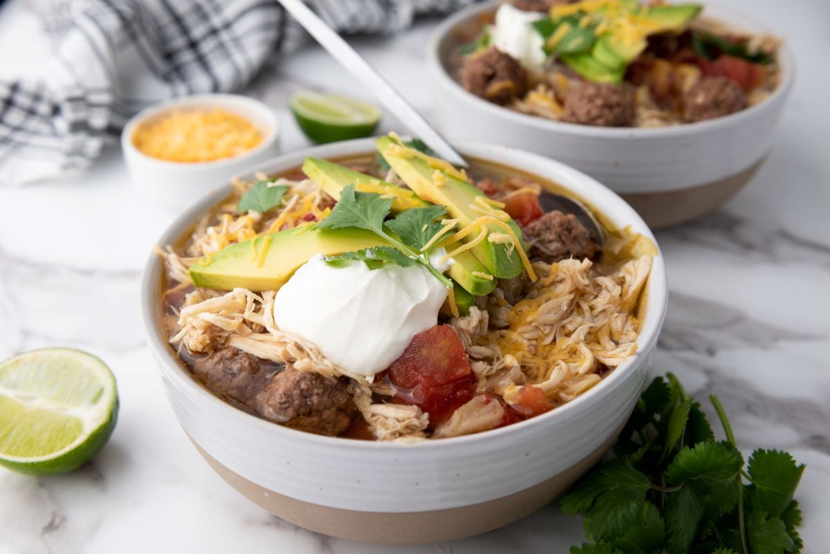 Two bowls of chili topped with avocado slices, sour cream, shredded cheese, and cilantro. Sliced lime and cheese bowl in the background on a marble surface.