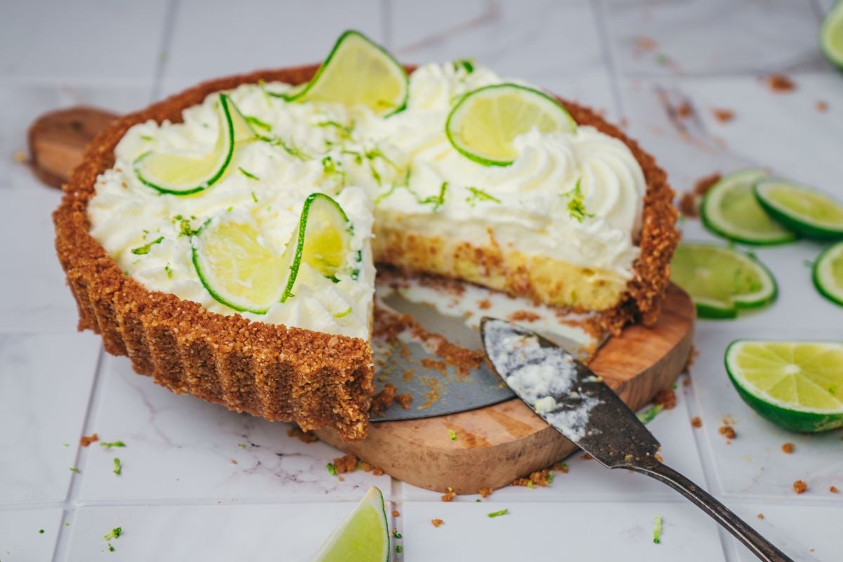 A key lime pie topped with whipped cream and lime slices sits on a wooden board. A slice has been cut and a pie server rests nearby, with crumbs and lime wedges scattered around.