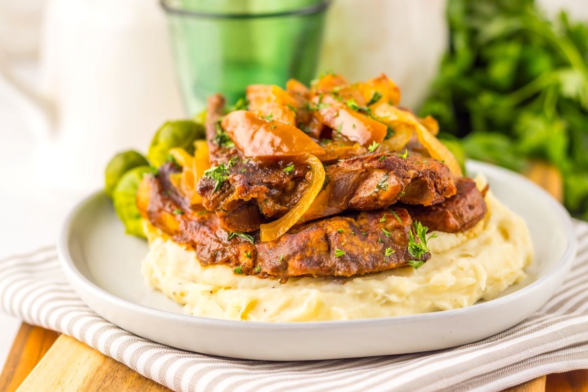 A plate of mashed potatoes topped with slices of cooked meat, sautéed onions, and herbs, served with Brussels sprouts.