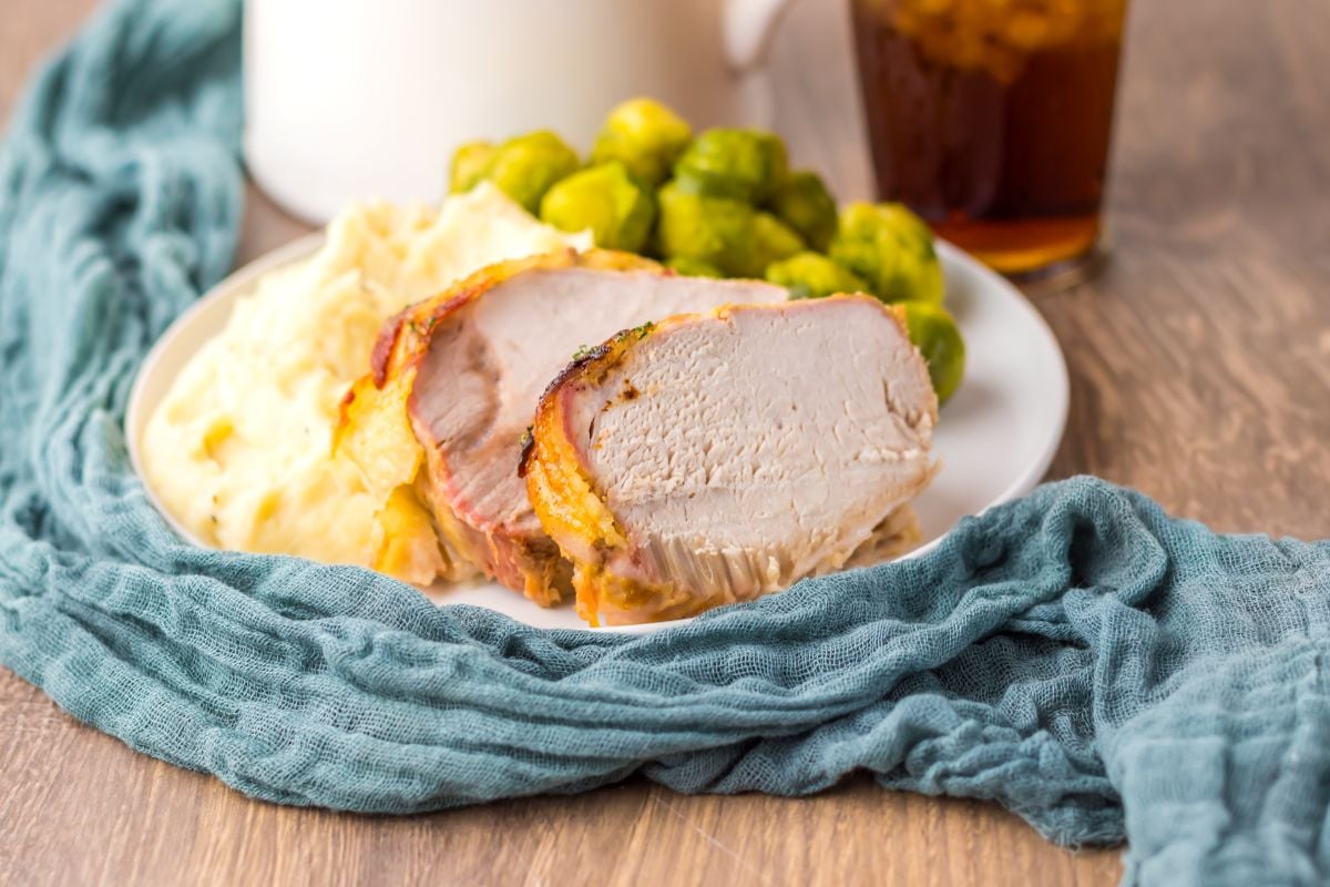 Plate with sliced roasted pork loin, mashed potatoes, and Brussels sprouts, set on a wooden table with a blue cloth and a glass of iced tea in the background.