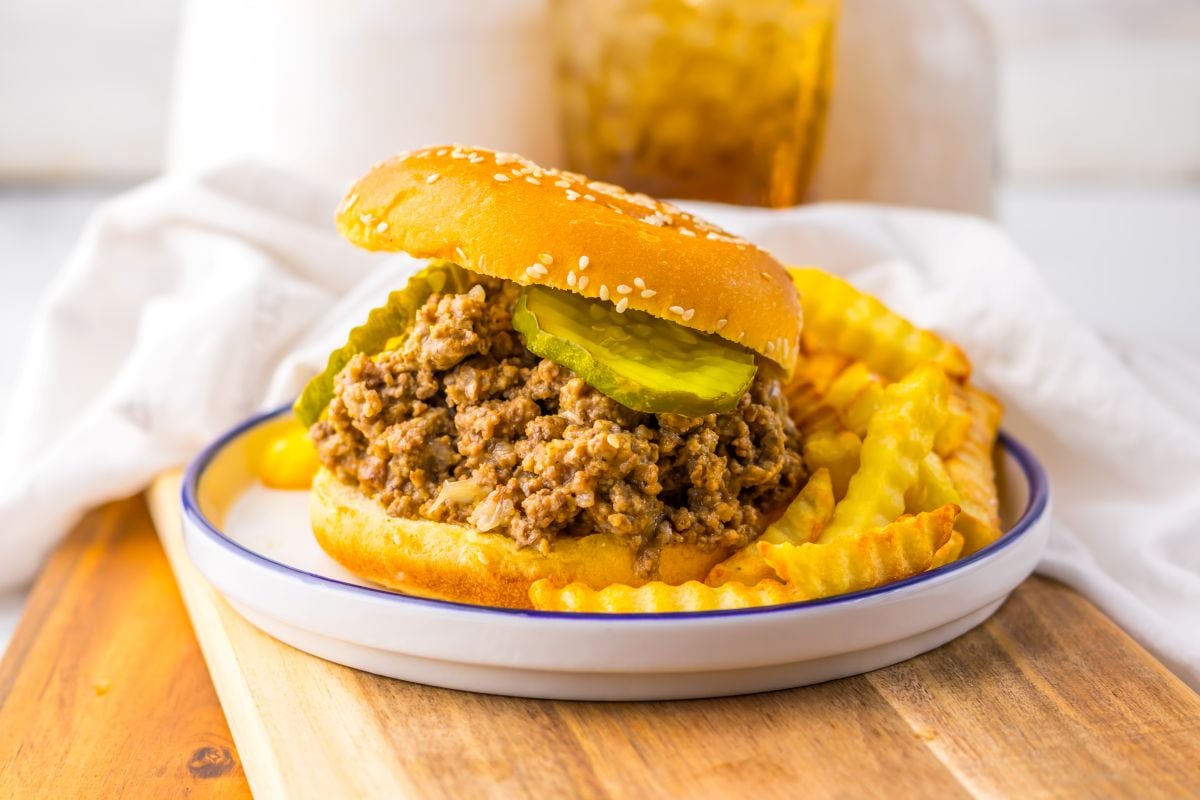 A sloppy joe sandwich with pickles on a sesame bun served with crinkle-cut French fries on a plate.