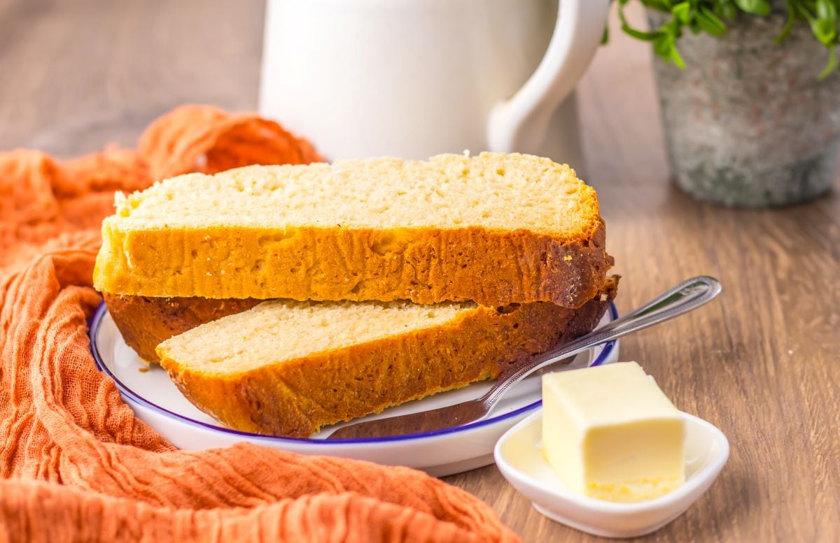 Two slices of bread on a plate with a butter knife, next to a small dish of butter, an orange cloth, and a white pitcher in the background.