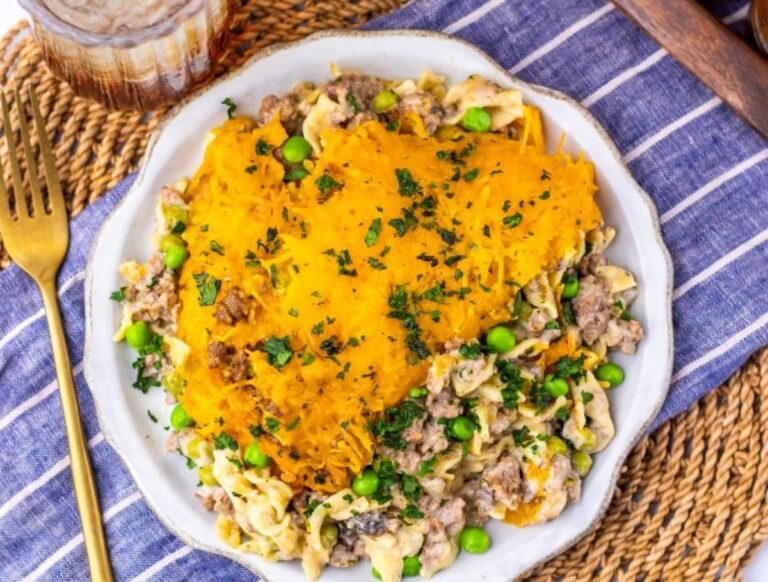 A plate of shepherd’s pie topped with mashed potatoes, ground meat, peas, and herbs, placed on a striped cloth next to a gold fork and a glass of beverage.