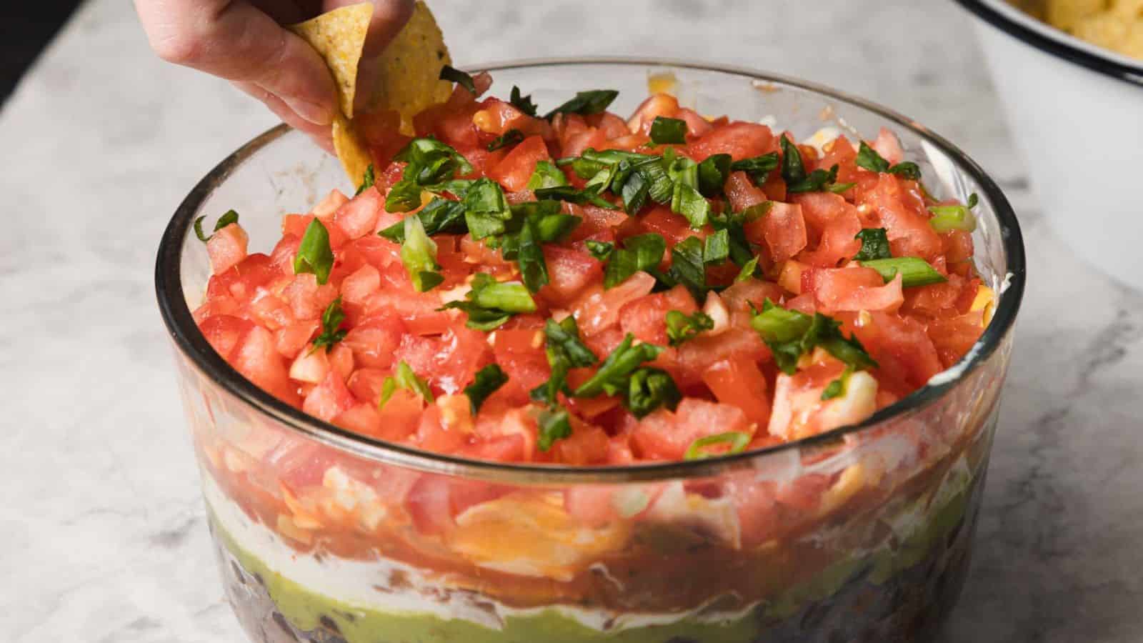 A hand dips a tortilla chip into a layered dip in a glass bowl. The dip consists of chopped tomatoes, green onions, cheese, beans, guacamole, and sour cream, arranged in visible layers. The bowl is on a marble surface, with more chips in the background.