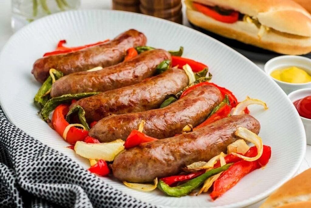 Five cooked sausages served on a white plate with sautéed red and green bell peppers and onions; condiments and a sausage roll appear in the background.