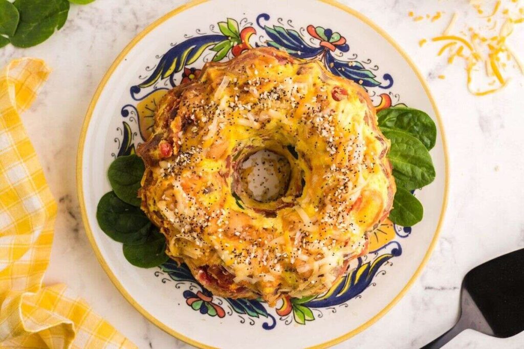 A baked bundt-shaped savory dish topped with shredded cheese and poppy seeds, served on a decorative plate with spinach leaves.