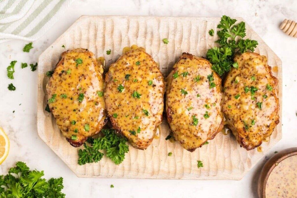 Four cooked pork chops topped with a grainy honey mustard glaze, garnished with chopped parsley, arranged on a light wooden serving board.