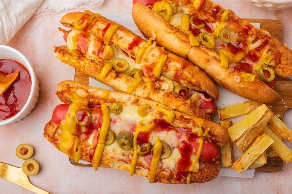 Three hot dogs topped with melted cheese, sliced green olives, mustard, and ketchup, served with a side of French fries on a wooden board.