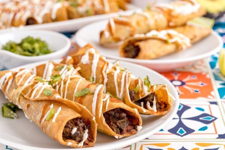 Three beef-filled taquitos are arranged on a white plate, topped with a drizzle of sour cream and chopped cilantro, with more taquitos and garnish in the background.