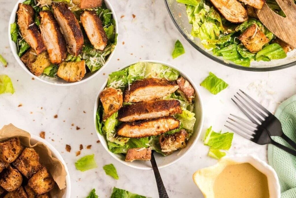 Bowls of Caesar salad topped with grilled chicken slices and croutons are arranged on a marble surface, with dressing, extra croutons, and utensils nearby.
