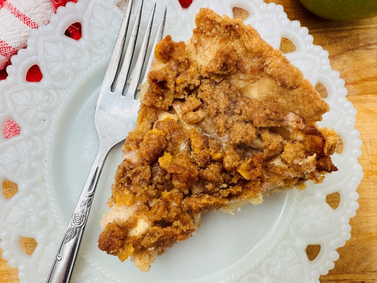 A slice of crumb-topped pie on a white decorative plate with a fork next to it. A red and white cloth is partially visible in the corner.