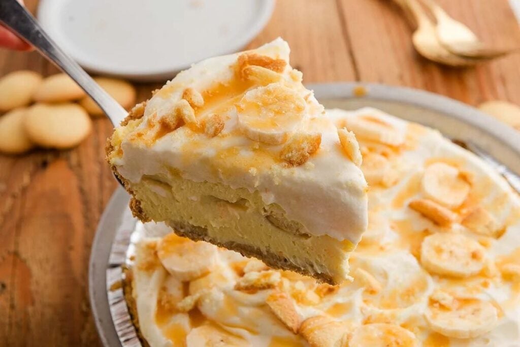 A slice of banana cream pie with whipped topping and cookie pieces is lifted from a pie tin on a wooden table.