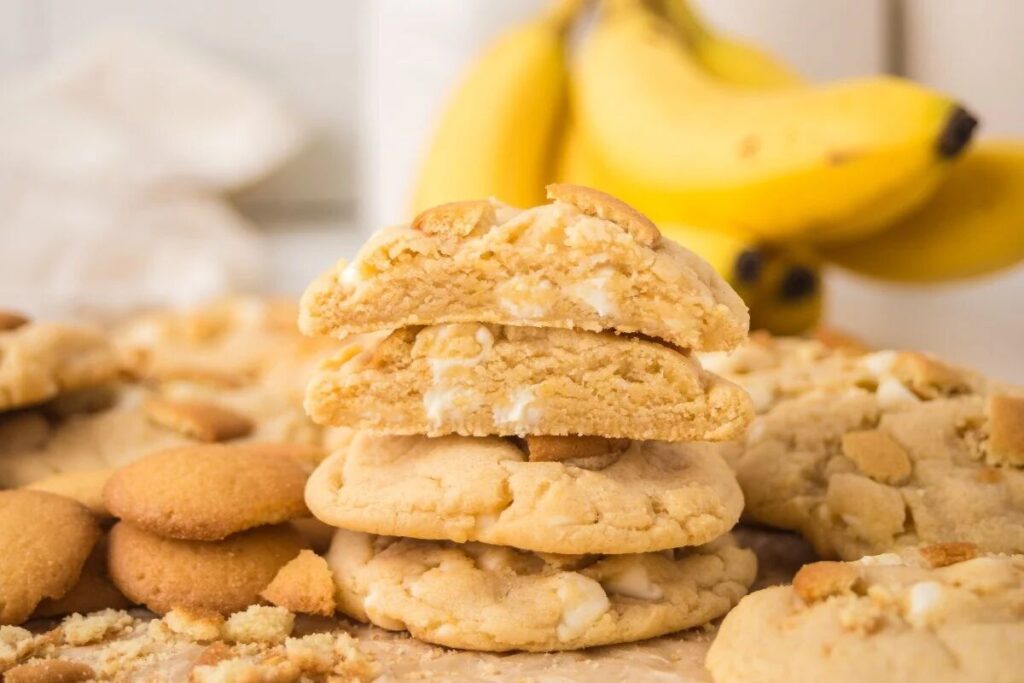 A stack of cookies with a broken cookie on top showing a soft, creamy center; bananas are blurred in the background.