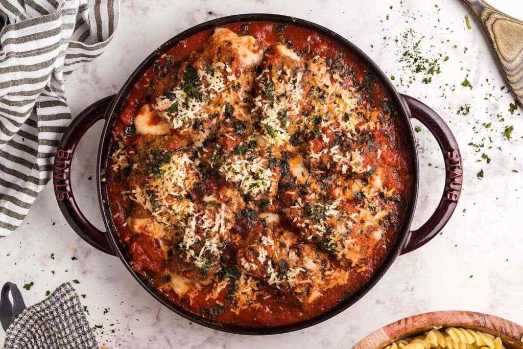 Round baking dish filled with baked chicken parmesan in marinara sauce, topped with melted cheese and herbs, on a marble surface next to a striped towel.