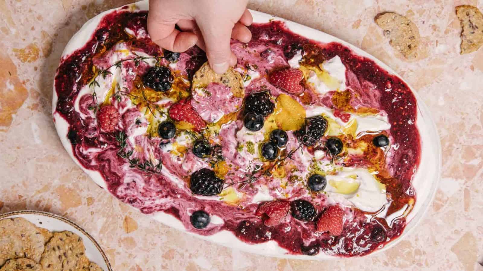A hand dips a cracker into a platter of whipped cheese topped with a berry sauce, fresh berries, herbs, and olive oil, set on a beige marble surface. A plate with more crackers is visible in the corner.