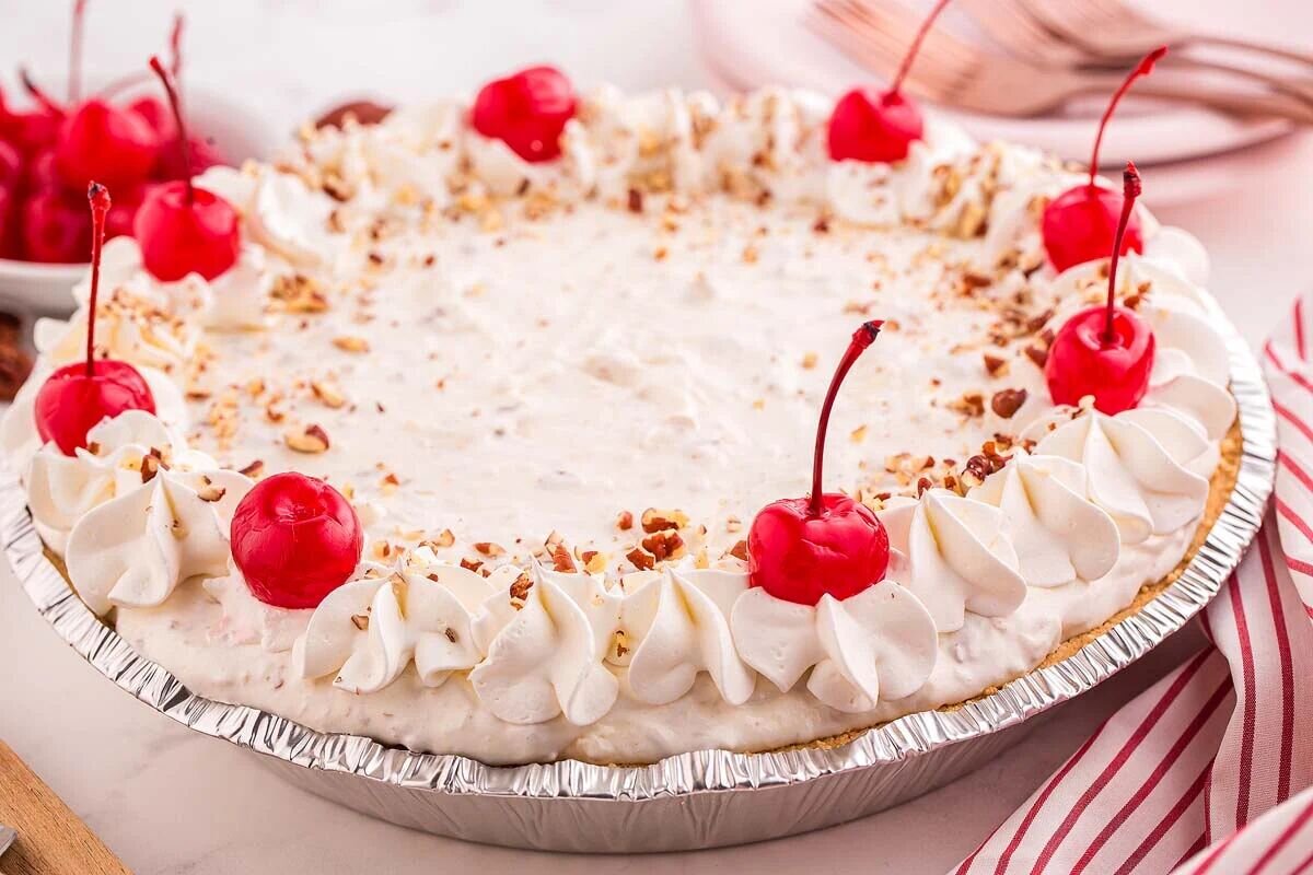 A cream pie topped with whipped cream, red cherries, and chopped nuts in a foil pie tin.