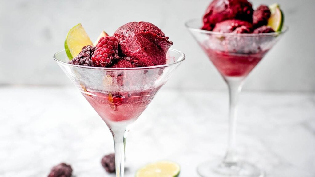 Two martini glasses filled with dark red berry sorbet, garnished with blackberries and a lime wedge, on a white surface.