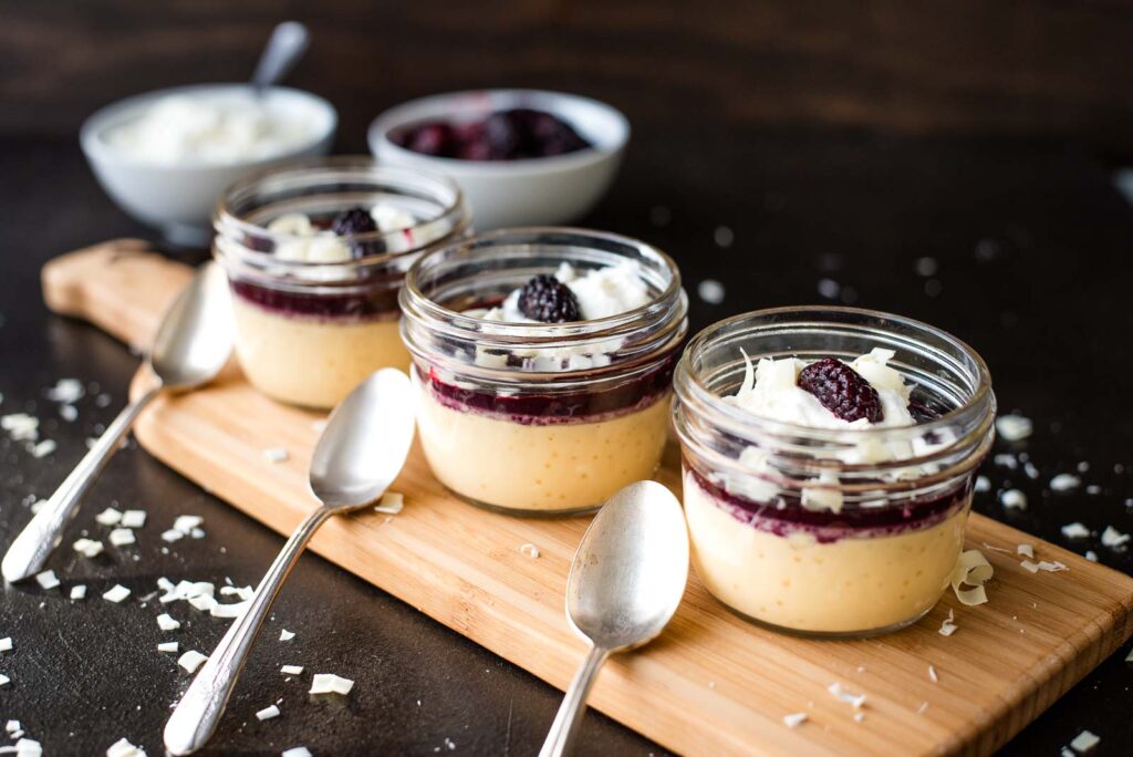 Three jars of layered vanilla pudding and berry compote topped with white chocolate shavings and blackberries, placed on a wooden board with three spoons.