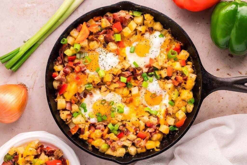 A cast iron skillet filled with a breakfast hash of eggs, diced potatoes, bell peppers, bacon, and green onions sits on a countertop with fresh vegetables nearby.