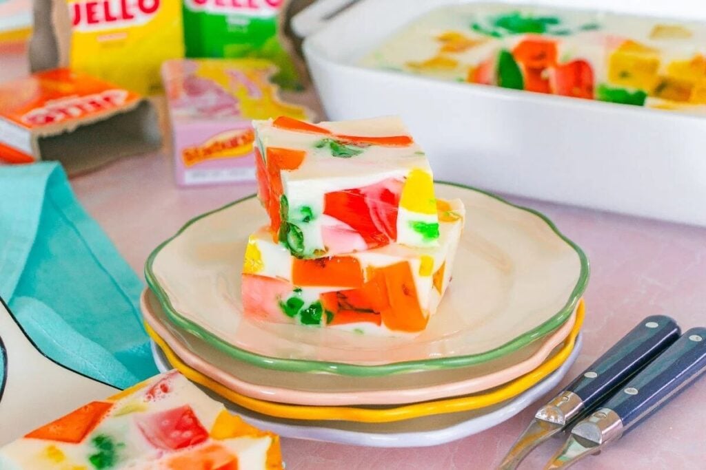 Three stacked slices of colorful stained glass Jello dessert on a plate, with boxes of Jello and a casserole dish in the background.