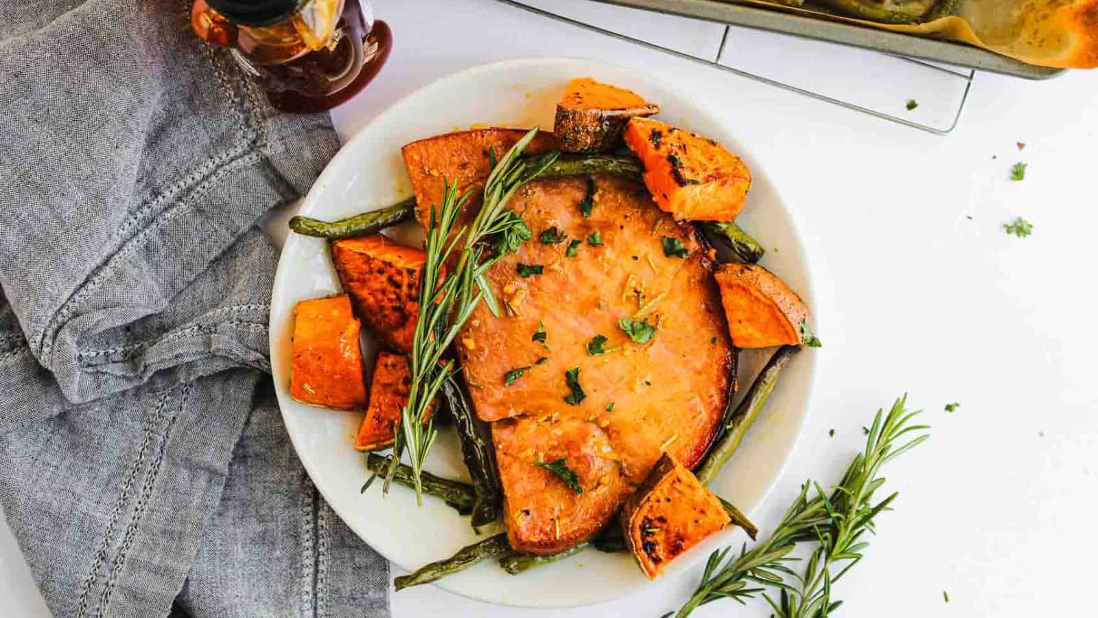 A white plate with a serving of glazed ham garnished with rosemary, surrounded by roasted sweet potato chunks and green beans. A gray napkin and a bottle of syrup are beside the plate.