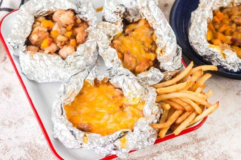 Four foil packets filled with cooked chicken and vegetables, some topped with melted cheese, served on a tray with a side of French fries.