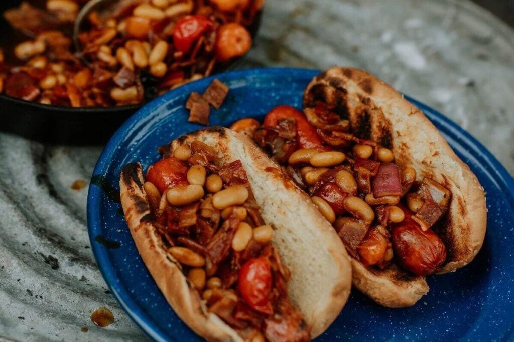 Two grilled hot dog buns filled with baked beans, chopped tomatoes, and small pieces of cooked bacon on a blue plate.