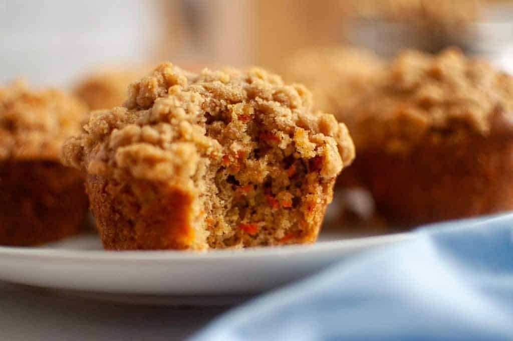 A close-up of a Carrot Muffin with Streusel Topping with a bite taken out, revealing a moist, spiced interior with visible pieces of carrot, on a white plate.